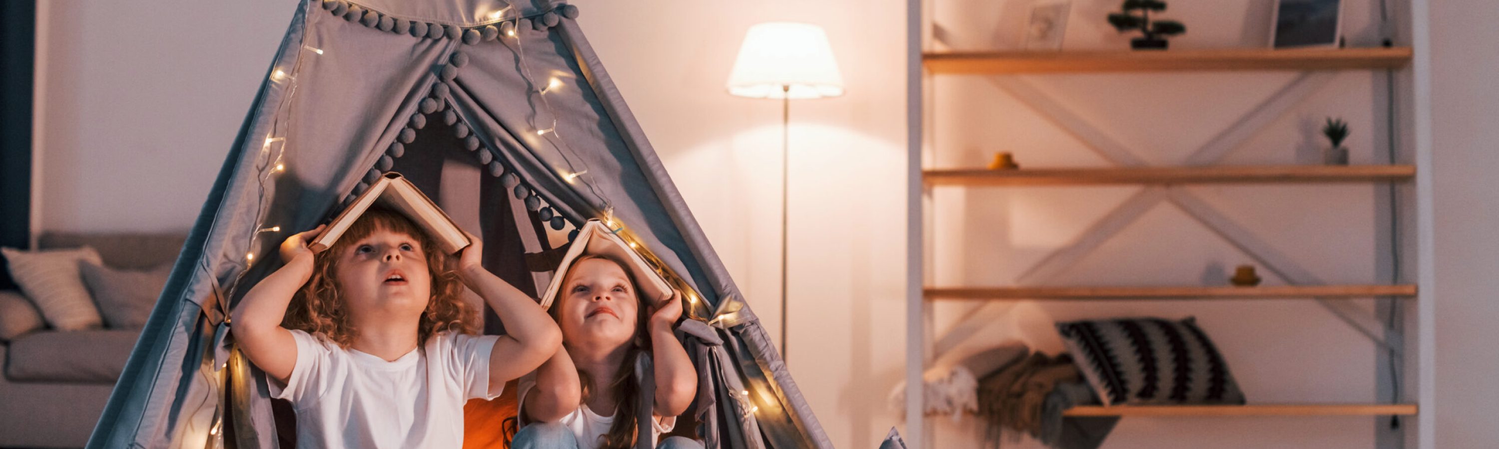 Holding books on head. Playful mood. Two little girls is in the tent in domestic room together.